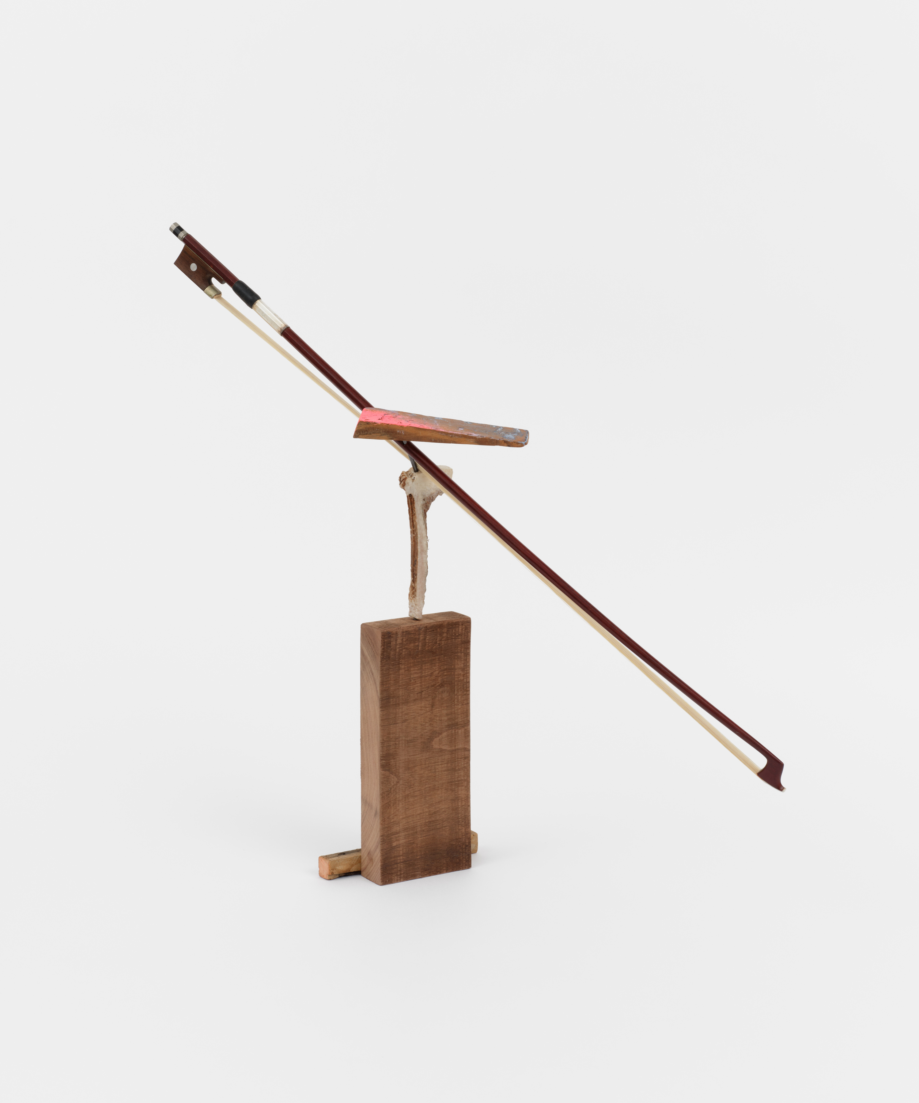 A free standing sculpture with a sliced piece of antler anchored into walnut wood. A bow used on string instruments balances on top of the antler with a triangular piece of wood brushed with pink paint. 