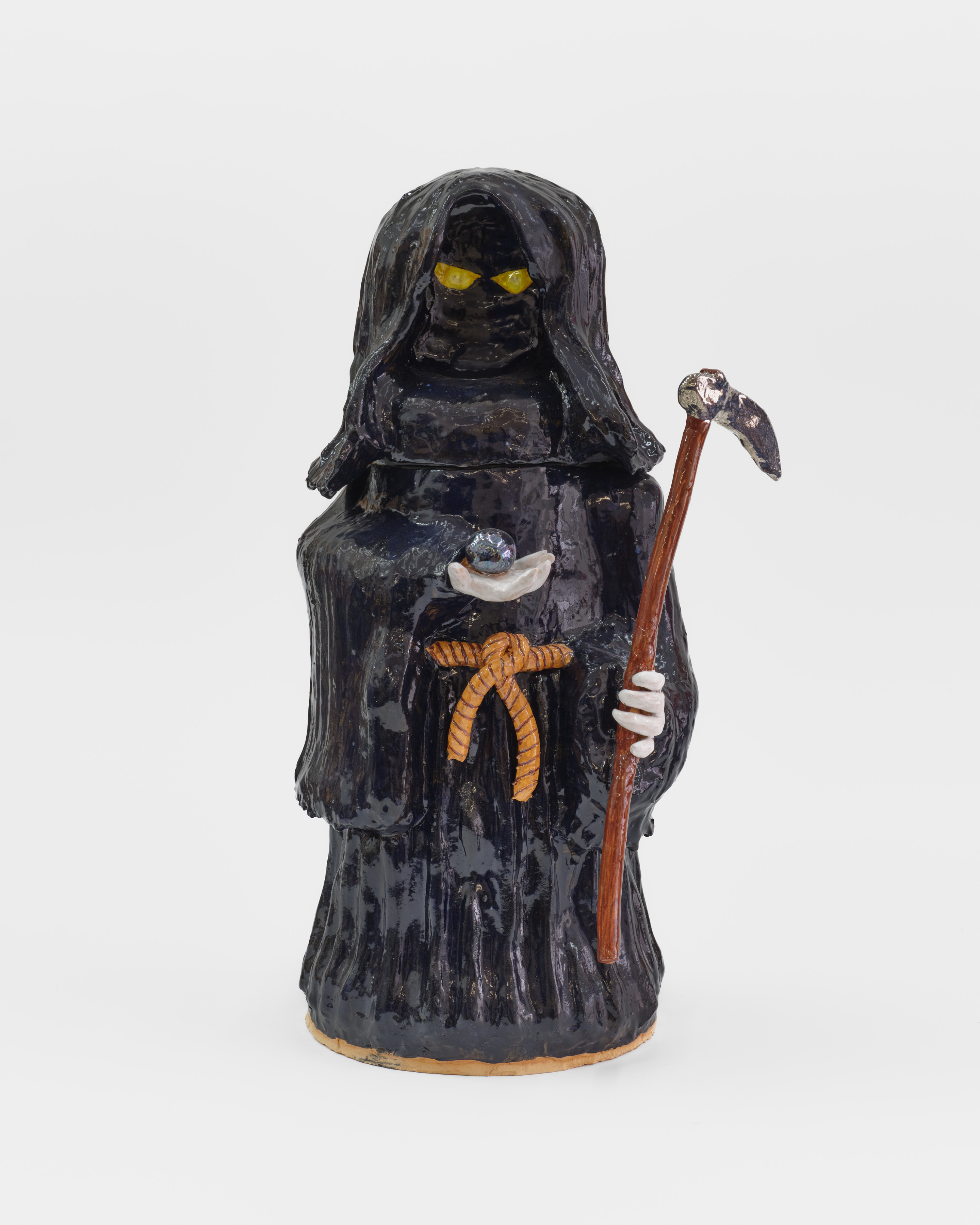 A ceramic sculpture of a black cloaked figure carrying a scythe and holding a shining stone. 