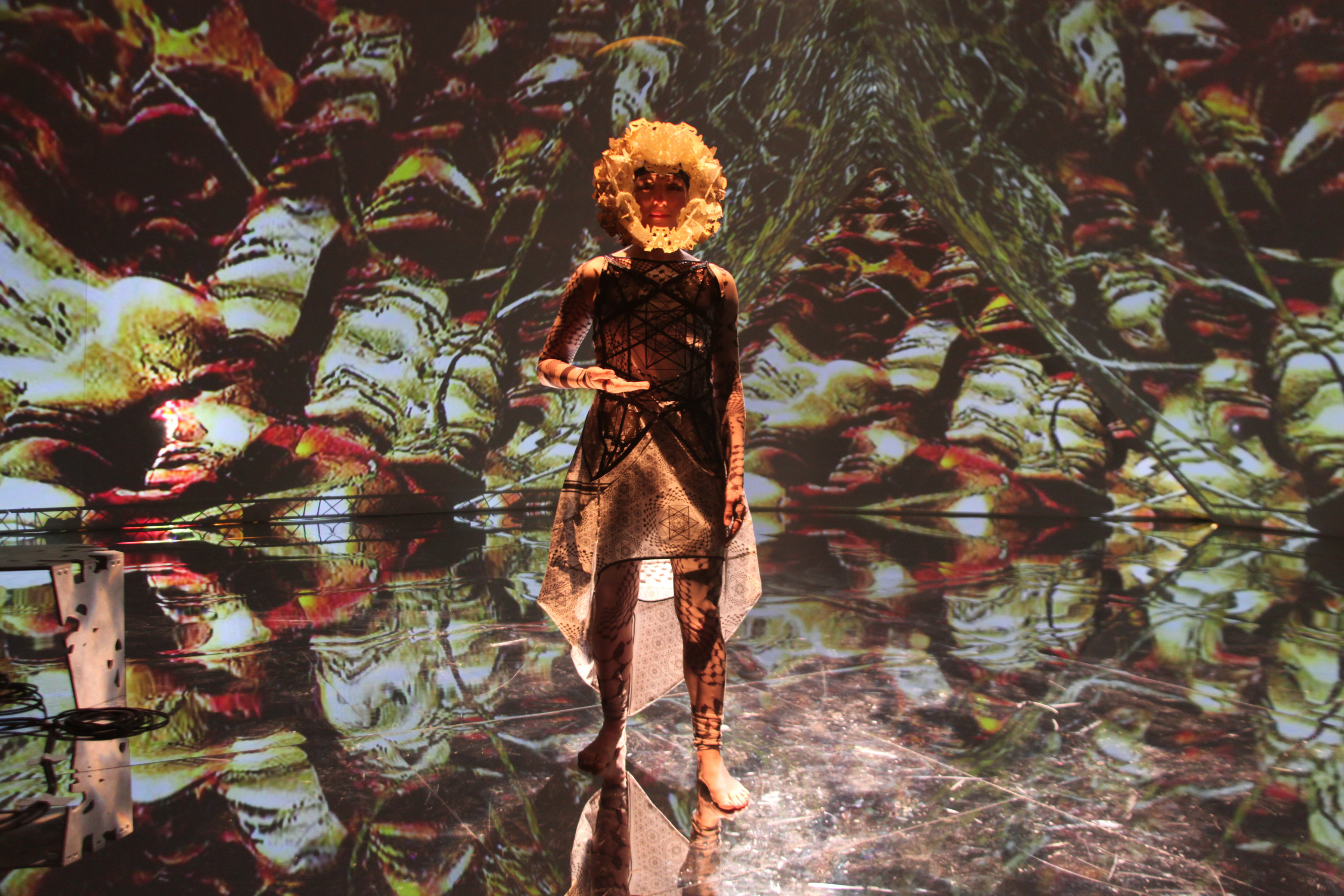 a woman wearing a mask staring directly at the camera; a psychedelic projection of color is displayed on the walls surrounding her