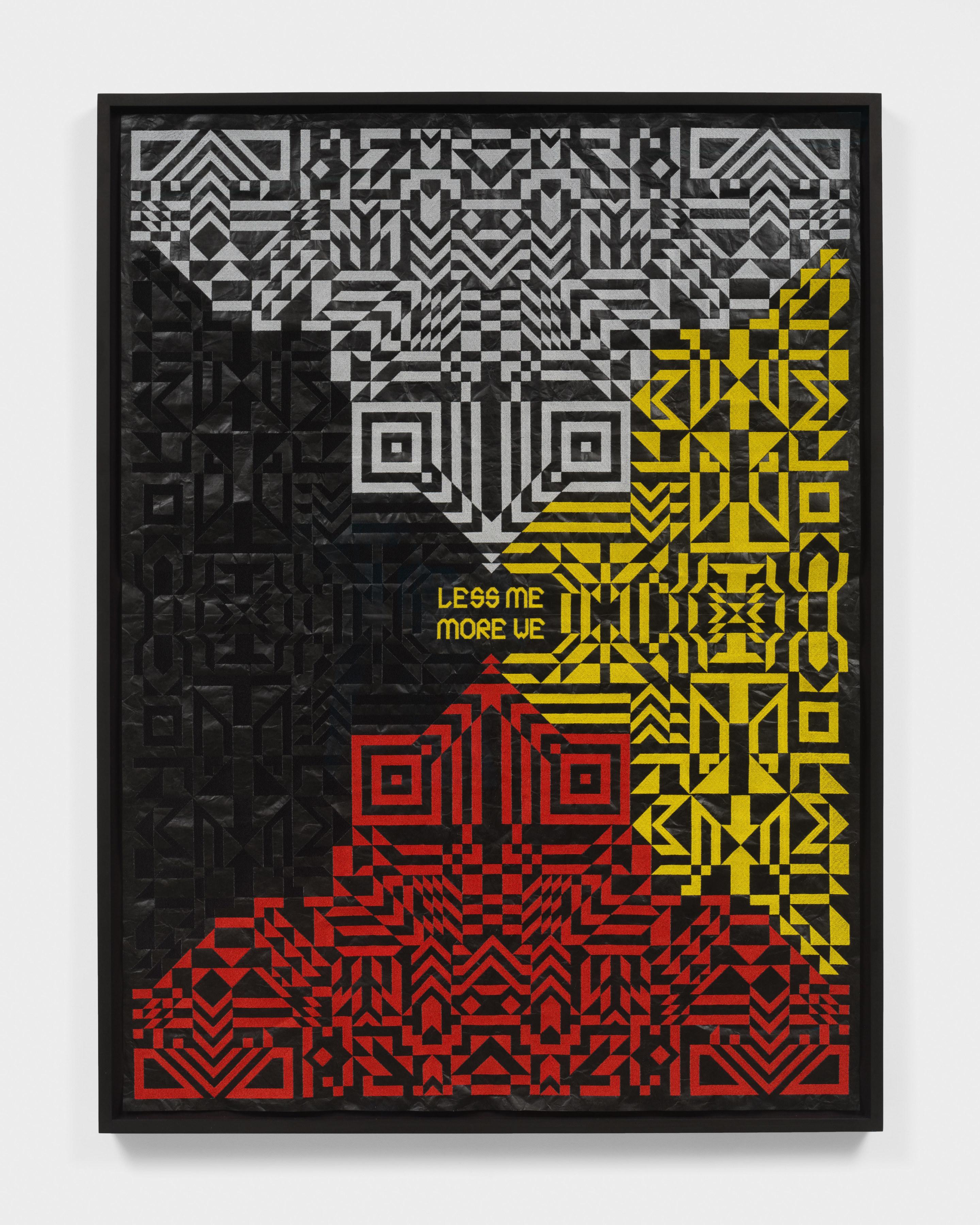 A framed artwork with red, white black and yellow embroidered patterns on black roofing material with the words "Less Me More We" in the center.