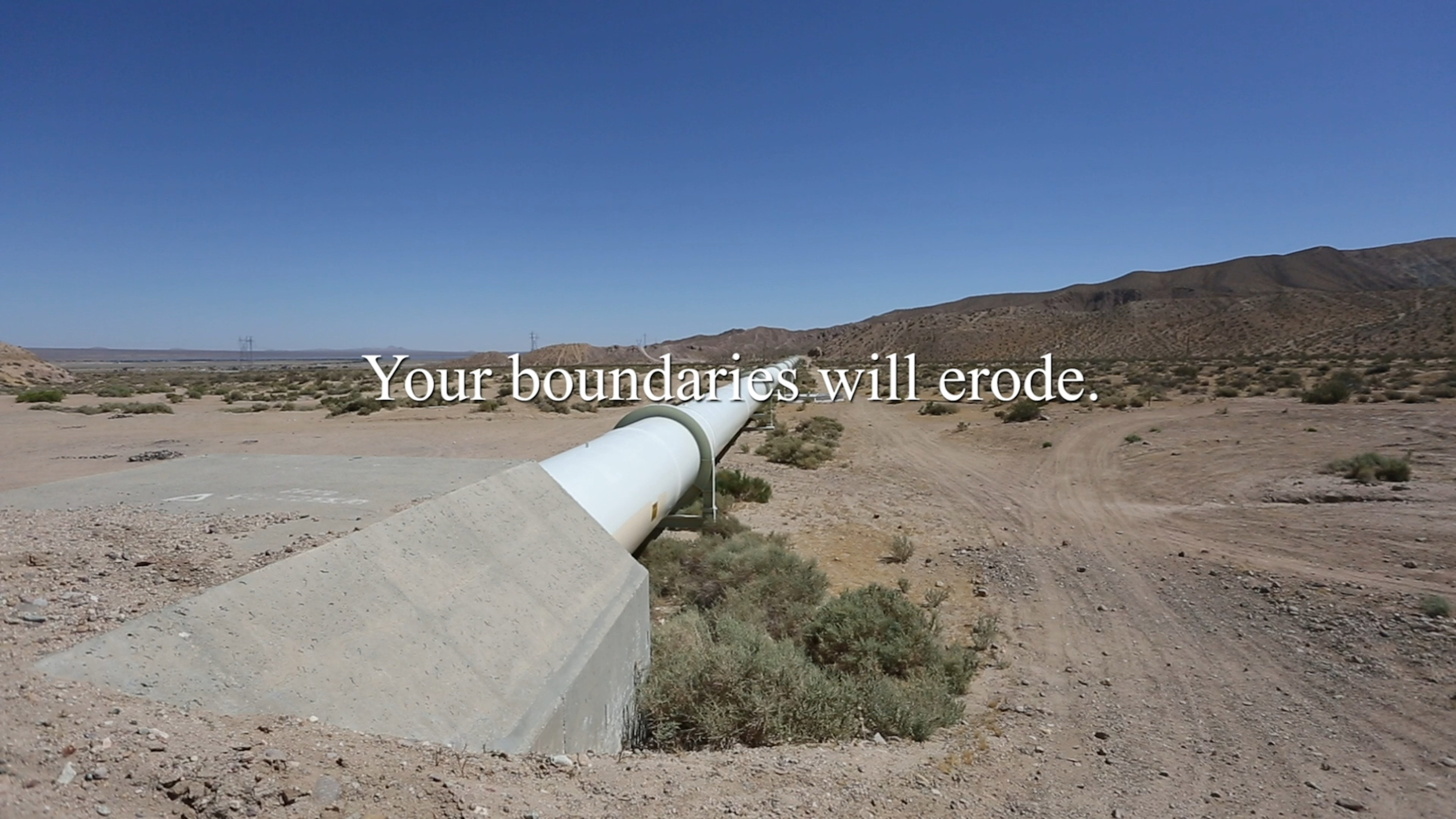 A film still of a pipeline across a desert landscape with the phrase "Your boundaries will erode" across the center.