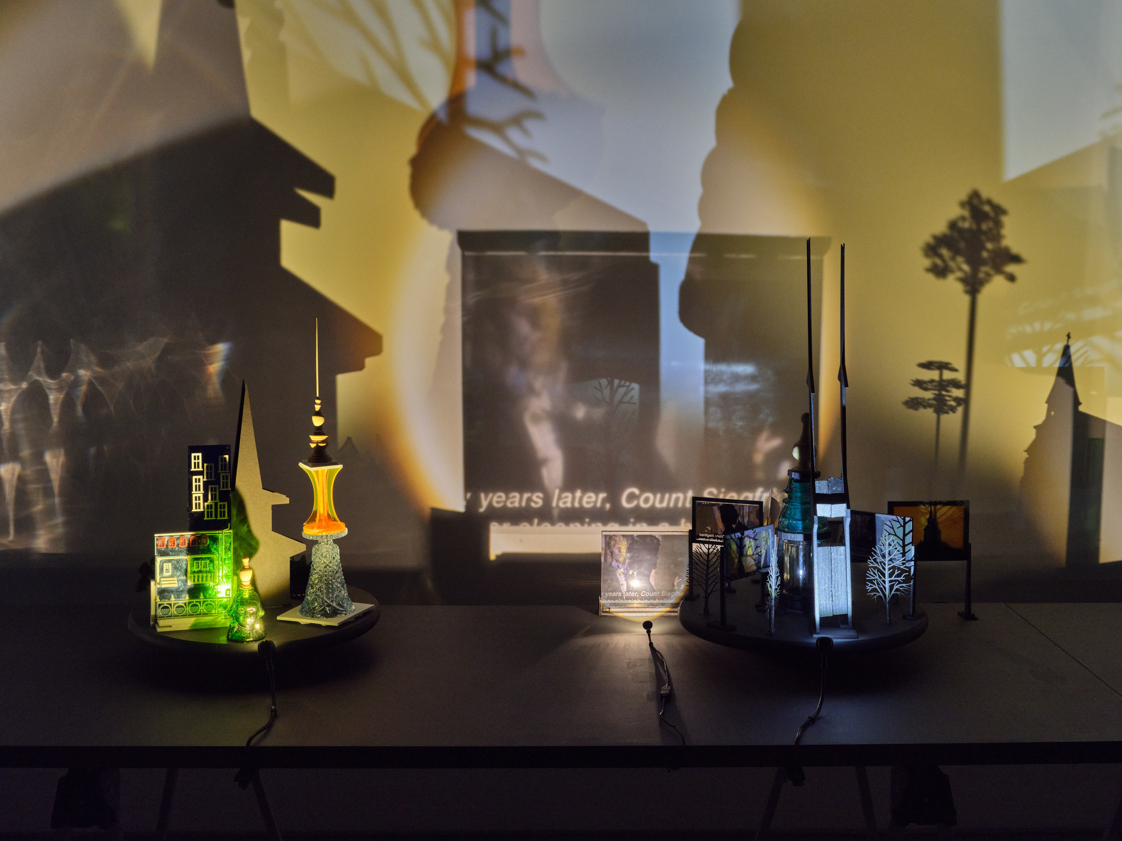 Two table top sculptures comprised of a miniature silhouettes of steeples, miniature trees, colored plexiglass, upside down crystal vessels and photographs printed on plexi that project onto the walls surrounding.