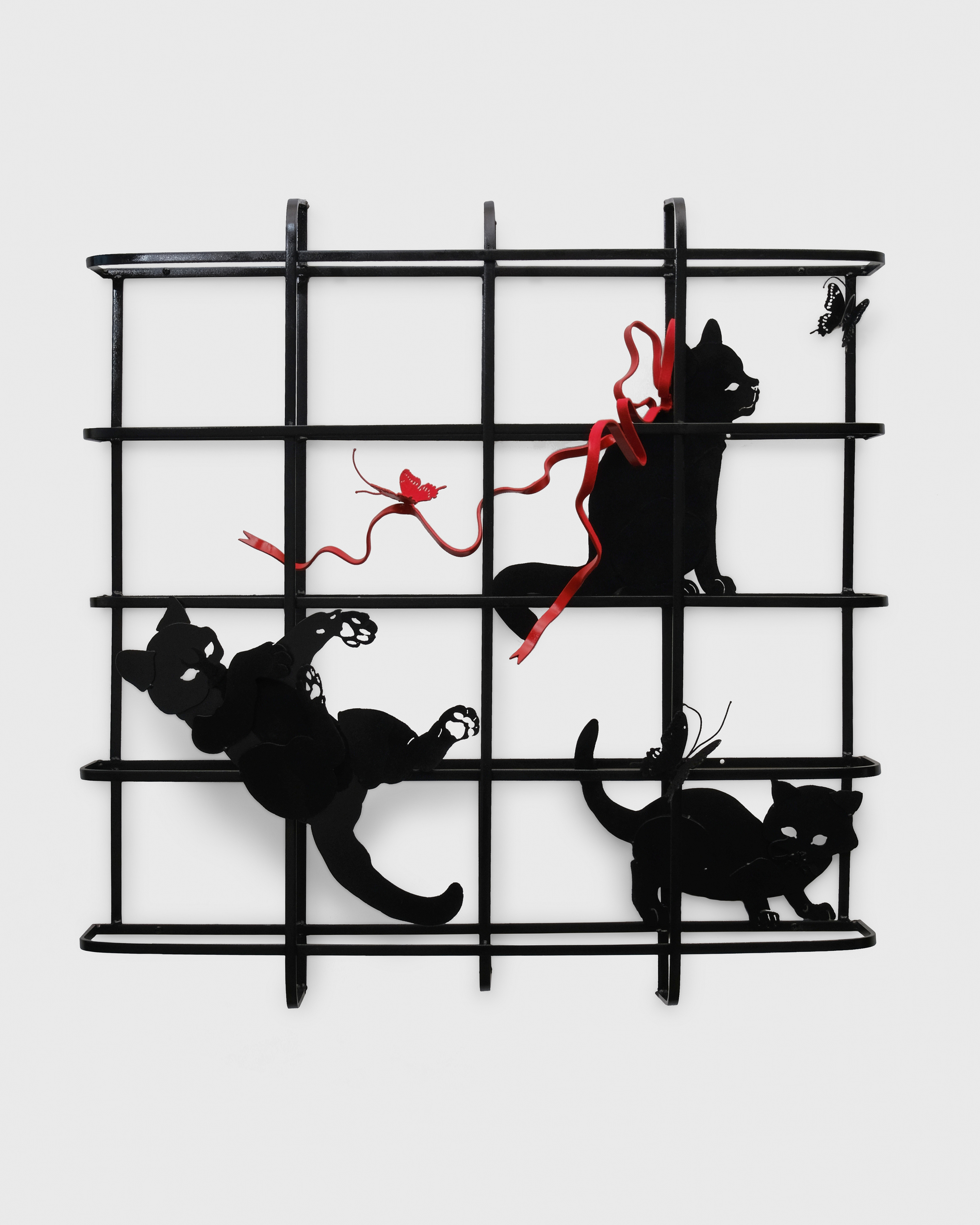 A wall mounted metal sculpture of three black cats within a grid like frame with a red ribbon woven through the grid with a butterfly atop.