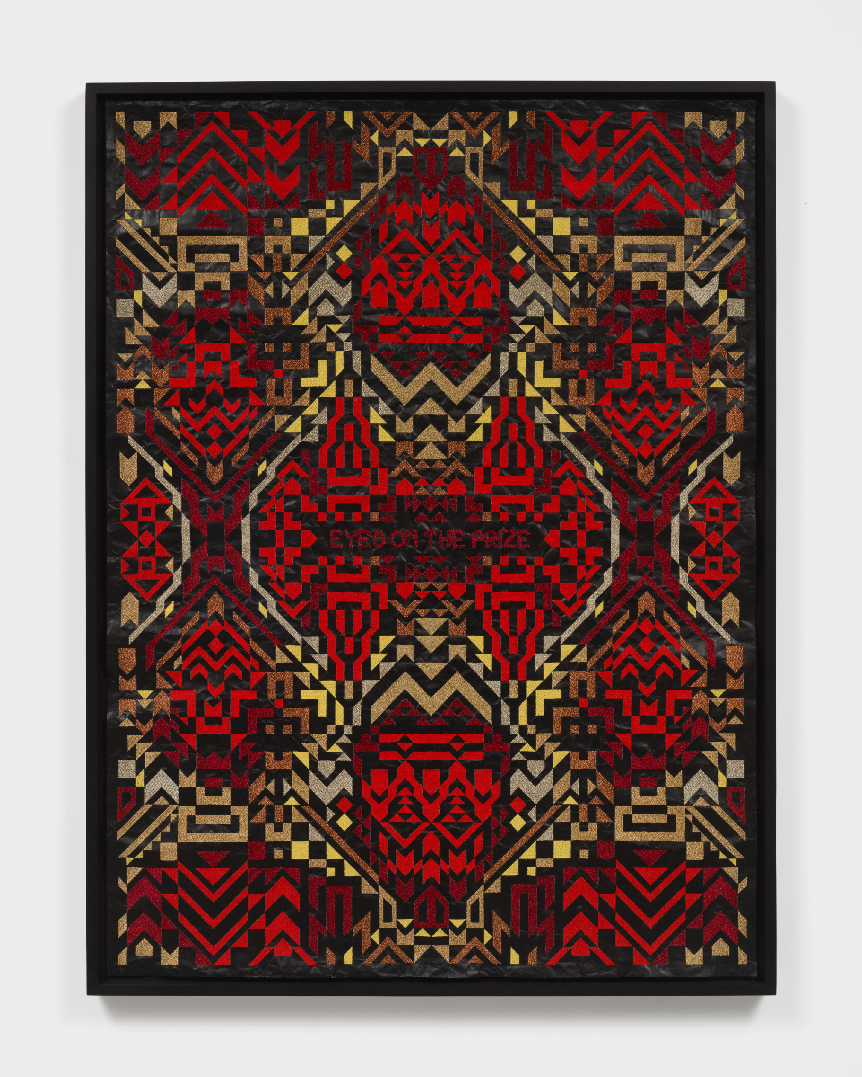 A framed artwork with red and yellow embroidered patterns on black roofing material with "Eyes on the Prize" in the center. 