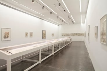 A gallery space with ink drawings installed on walls and a wooden display table placed at the center