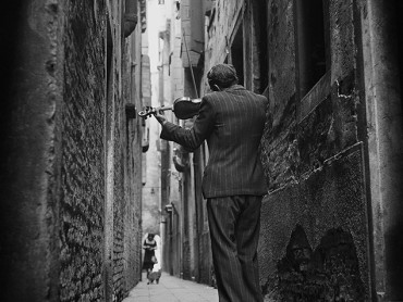 Black and white photo of a violinist playing in a tight alleyway where a woman and a cat walk in the distance