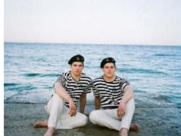 Two men sit in front of the sea, wearing berets and striped shirts.