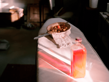 A stream of light illuminates a bright orange liquid in a glass bottle which is on an ironing board next to a bowl of cherries.