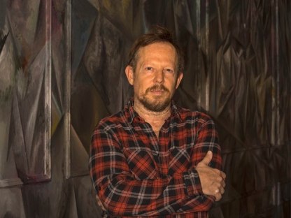portrait of Guillermo Kuitca standing with his arms crossed in front of a cubist-style abstract wall painting