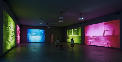 installation view of Mapping the Studio showing five colorful projections along three walls with two people sitting on stools in the middle of the room