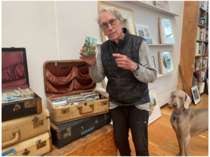 A man in glasses holding up a postcard with a grey dog next to him and old suitcases, and artwork in the background