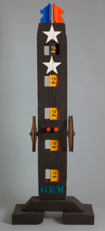 A 71 by 15 1/2 by 15 1/2 inch sculpture consisting of a wooden beam on a wooden base. The wood has been painted black, and the work's title, "Gem," painted in blue stenciled letters across the bottom. Five squares run down the front of the work; half the square has been hollowed other has been painted white with a black number two, except for the top square which is painted black with a white two. Two red pegs have been affixed between the second and third squares, and an iron wheel has been affixed to the left and right sides of the sculpture next to the pegs. A white star has been painted above and below the stop square, and on top of the sculpture are two wooden pieces resembling a crest, one blue and the other red.