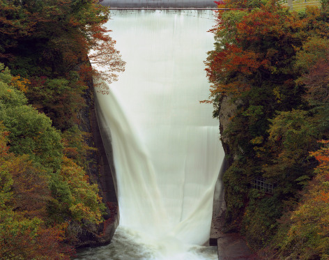 Color long exposure phto of water falling over a dam in a forested area during autumn.