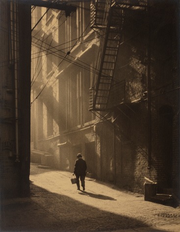 Black and white image: shaft of morning light illuminates person carrying back down a city alleyway.