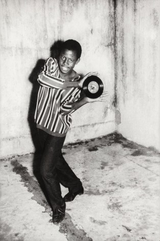 Black and white photo of a Black man posing dramatically in a striped shirt with a 45 inch record.