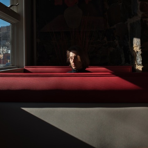 Woman sitting alone in a diner booth, next to a window, in slanting light. She is looking at the camera.