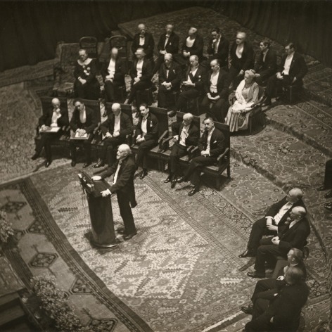 Black and white photo of the 1929 Nobel Prize cerenmonty. A grey bearded man speaks at a podium, while a formally dressed audience sits behind him.