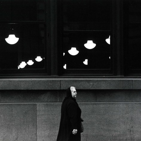 Black and white photos of a woman walking awn sidewalk, with a window showing midcentury pendant lights behind her—the lights are surrounded by blackness.