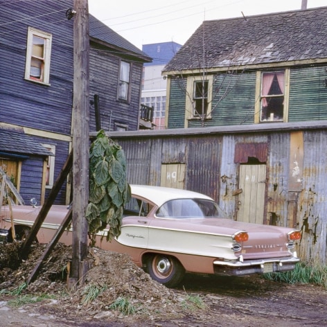Color photo of a new 1950s era Pontiac parked in a dirt driveway in front of dilapidated buildings.