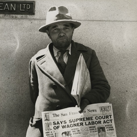 Black and white photo of a newspaper dealer with a copy of the San Francisco News from 1937 announcing Supreme Court decision on Wagner Labor Act
