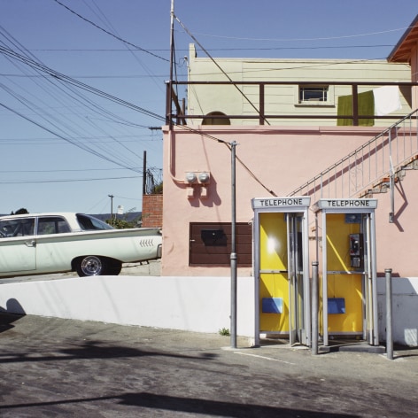 Color photograph of two yellow phone booths wit ha pink wall and a light green 1960s car behind them.