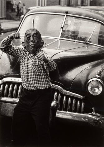 Black and white photo of a young boy posing like a boxer in front of a car while wearing a rubber mask.