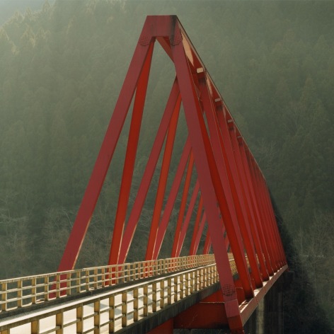 Photo of a red truss bridge in misty and slanting afternoon light with a wooded mountain behind it.