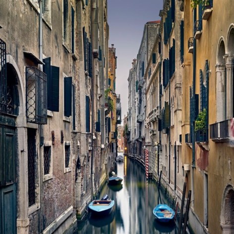 Long exposure color photograph of a side can in Venice Italy at dusk. Three small blue boats are docked at the sides of the canal.