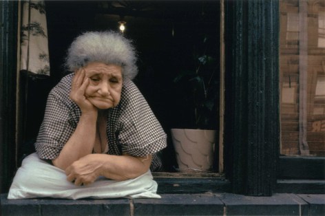 Color photograph of an old woman leaning on a window sill frowning.