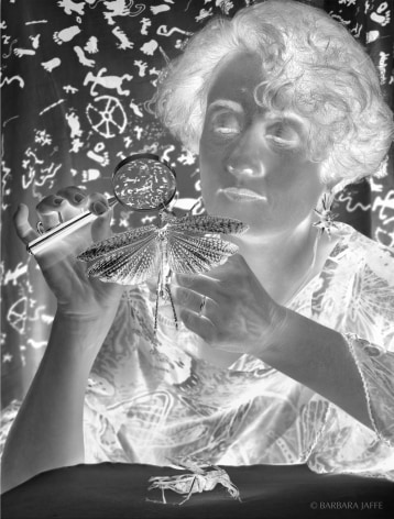 Black and white negative photograph of woman holding and examining a large winged bug under a magnifying glass.