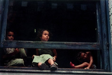 Color photo of three kids sitting in an open window sill, peering out.