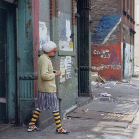 Color photo of an older woman in blue and yellow striped socks with a colorful mismatched outfit walking on a grimy street with graffiti in New York City.