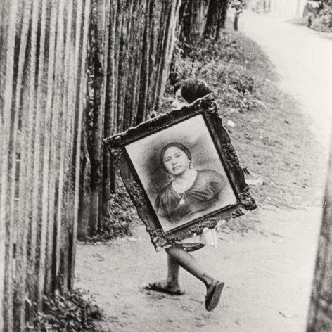 Black and white photo of a young girl carrying a large framed portrait of woman.