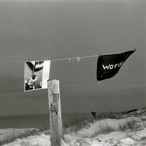 Black and white photo of a drying line with a photo and a piece of paper inscribed with "words", blowing in the wind in front of a wintry seascape.