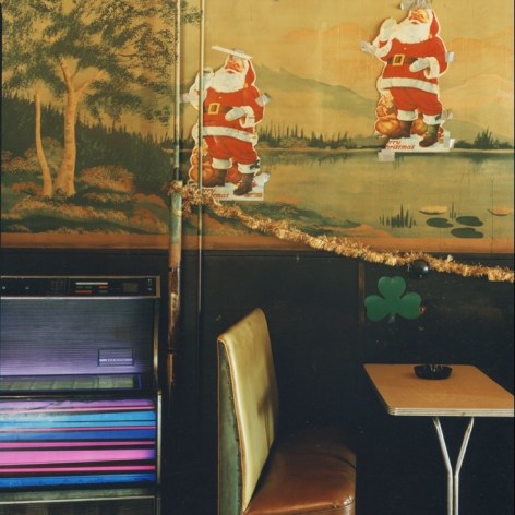 Color photo of oa booth with an ash tryain a working class bar, a color jukebox on the left. The wall is decorated with a faded landsca[e print, with holiday deceptions (two Santas and a shamrock) over top.