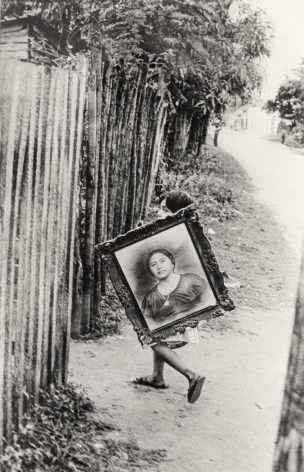 Black and white photo of a young girl nearly hidden behind a framed portrait of a woman that she's about to carry behind a fence.