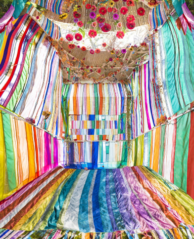 Photo of a sun filled Sukkah (tent) made form brightly colored fabric, with flowers hanging from the ceiling.