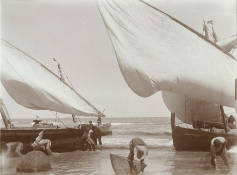 Black and white photo of young men wading alongside boats while working fishing baskets in the water.