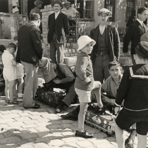 Black and white photo showing a young boy on a cobblestone street, among a group of people getting shoeshines.