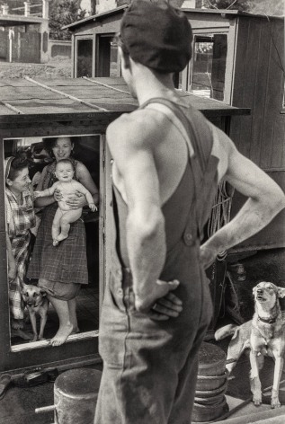 Black and whote hoto of a min in overalls with hand on his hips with back to the camera looking at a house boat, two omen o=are on the boat, the younger holds a baby, there are two dogs.