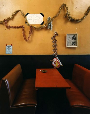 Color photo of a diner booth, red seats with a gold wall, and haphazardly attached tinsel and Xmas decorations.