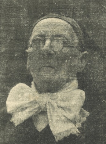 Black and white portrait of an old man wearing a cap, a large neck kerchief, and round glasses.