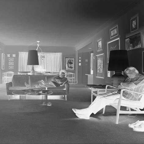 Negative printed photo of a couple sitting in a midcentury living room with the TV on. The man sleeps in a char while the woman looks at the camera.