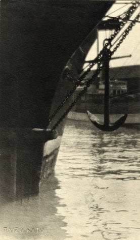 Black and white photo of ship's anchor hanging over the water at a port.