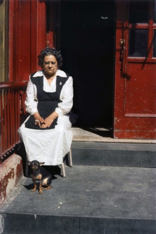Color photo of an old woman on a stoop with a small dog, with a red door behind them.