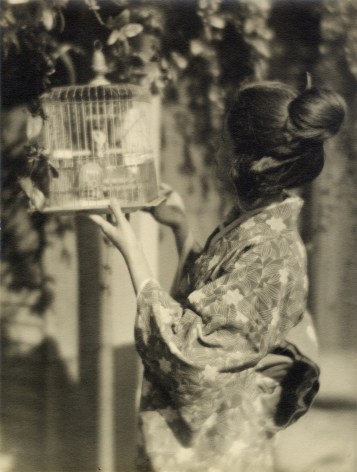 Japanese American woman in a kimono hold. birdcage and peers in at the bird inside.