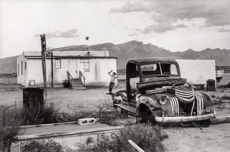Black and white photo of a shirtless boy shooting a basketball on a desolate desert lot that holds a mobile home and and abandoned vintage car.