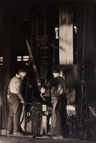 Black and white image: Workers drop forging steel components in a factory.