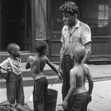 Black and white photo of an 1940s New York City street scene showing three young black boys standing in front of muscular white laborer.