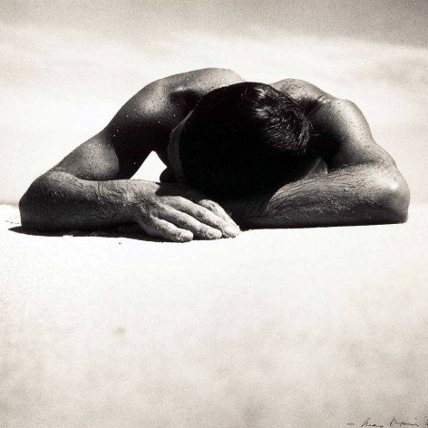 Black and white photo of a wet shirtless man sunbathing on a beach.